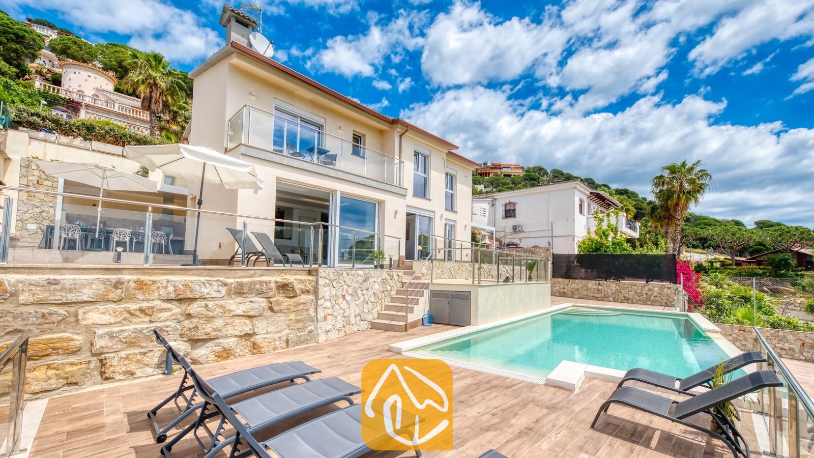Vakantievilla's Costa Brava Spanje - Villa BellaVista - Privézwembad bij Villa BellaVista met grijze ligbedden op een houten terras, glazen balustrades en een crèmekleurige gevel met stenen accenten en witte parasols.