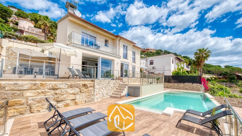 Vakantievilla's Costa Brava Spanje - Villa BellaVista - Privézwembad bij Villa BellaVista met grijze ligbedden op een houten terras, glazen balustrades en een crèmekleurige gevel met stenen accenten en witte parasols.