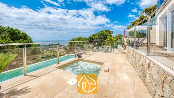 Vakantievilla's Costa Brava Spanje - Villa BellaVista - Een bruisende hot tub en zwembad op een stenen terras bij Villa BellaVista. Glazen balustrades bieden uitzicht op de kustlijn en de bomen onder een bewolkte, blauwe lucht.