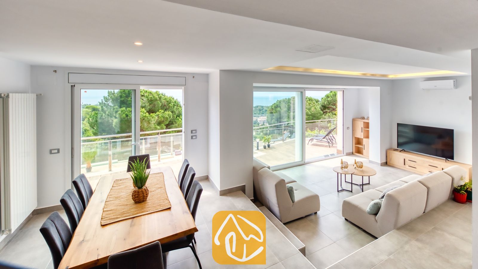 Vakantievilla's Costa Brava Spanje - Villa BellaVista - De open woon- en eetkamer van Villa BellaVista is voorzien van een houten tafel en grijze modulaire banken, met grote glazen deuren die uitkomen op een terras met uitzicht op zee.