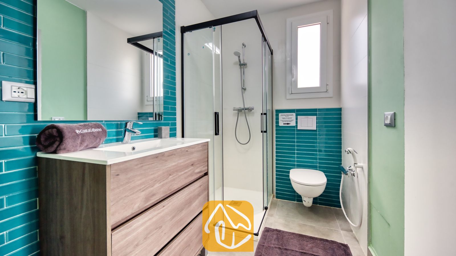 Vakantievilla's Costa Brava Spanje - Villa BellaVista - Moderne badkamer met teal-kleurige metrotegels, een wastafelmeubel met houtnerf en een glazen douchecabine met een zwart frame. Een hangend toilet en een paarse badmat zijn zichtbaar.