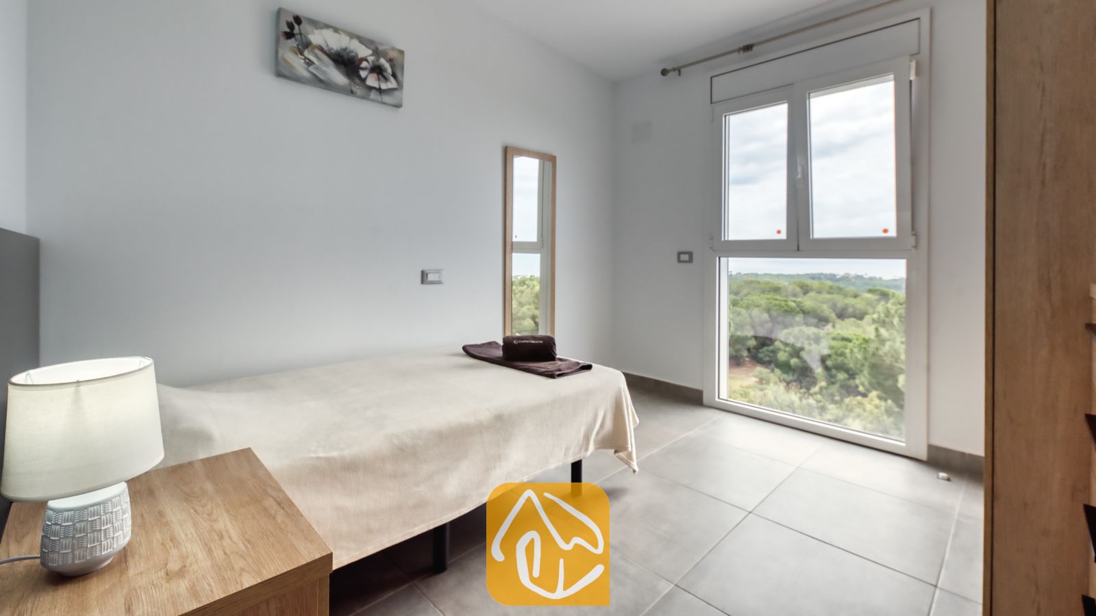 Vakantievilla's Costa Brava Spanje - Villa BellaVista - Eenpersoonskamer in Villa BellaVista, voorzien van een bed met crèmekleurig beddengoed, een houten nachtkastje met leeslamp en een groot raam met uitzicht op een weelderig groen bos.