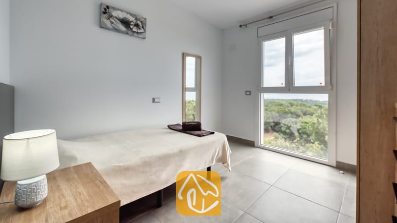 Vakantievilla's Costa Brava Spanje - Villa BellaVista - Eenpersoonskamer in Villa BellaVista, voorzien van een bed met crèmekleurig beddengoed, een houten nachtkastje met leeslamp en een groot raam met uitzicht op een weelderig groen bos.