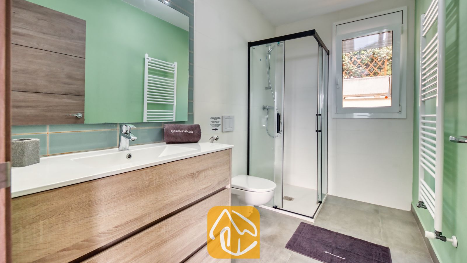 Vakantievilla's Costa Brava Spanje - Villa BellaVista - Moderne badkamer in Villa BellaVista, voorzien van een wastafelmeubel in licht hout, een glazen hoekdouche en muren in saliegroen.