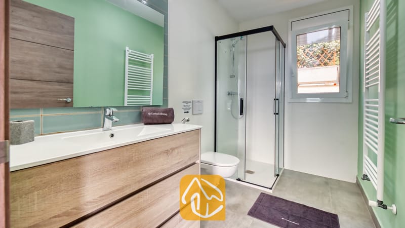 Vakantievilla's Costa Brava Spanje - Villa BellaVista - Moderne badkamer in Villa BellaVista, voorzien van een wastafelmeubel in licht hout, een glazen hoekdouche en muren in saliegroen.