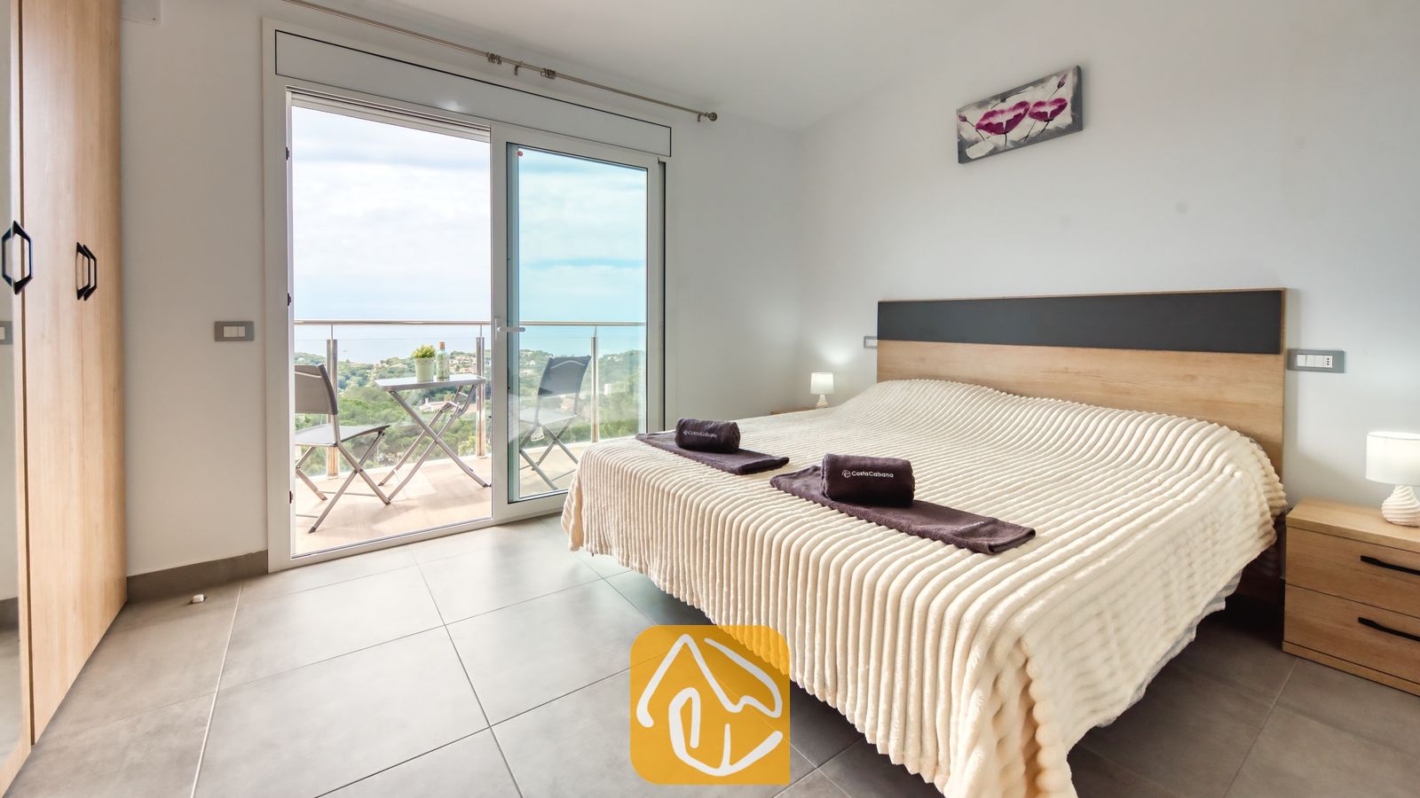 Vakantievilla's Costa Brava Spanje - Villa BellaVista - Slaapkamer in Villa BellaVista met een groot bed, crèmekleurig beddengoed met textuur en glazen schuifdeuren die uitkomen op een balkon met uitzicht op de zee en de groene heuvels.
