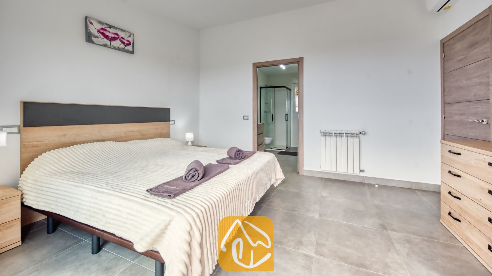 Vakantievilla's Costa Brava Spanje - Villa BellaVista - Tweepersoonsbed met paarse handdoeken in een slaapkamer voorzien van lichtgrijze tegels, houten meubilair en uitzicht op een aangrenzende badkamer met een glazen douche.