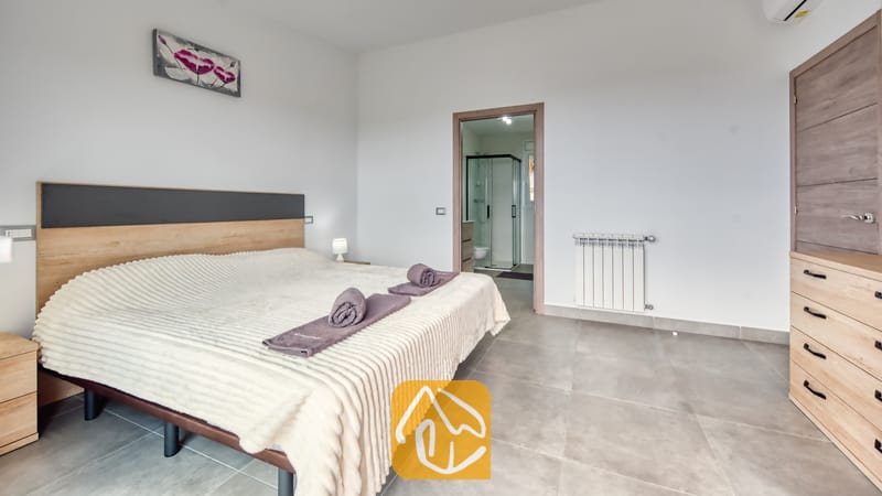 Vakantievilla's Costa Brava Spanje - Villa BellaVista - Tweepersoonsbed met paarse handdoeken in een slaapkamer voorzien van lichtgrijze tegels, houten meubilair en uitzicht op een aangrenzende badkamer met een glazen douche.