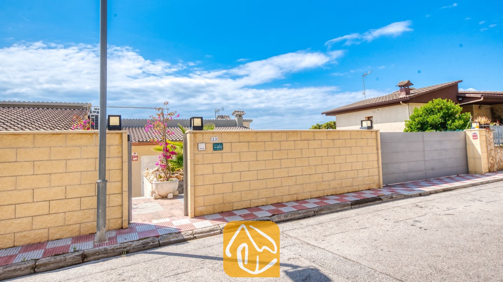 Vakantievilla's Costa Brava Spanje - Villa BellaVista - De geelstenen omheiningsmuur van Villa BellaVista, voorzien van een grijze schuifpoort, een open entree met roze bougainville en een rood-wit geblokt trottoir.