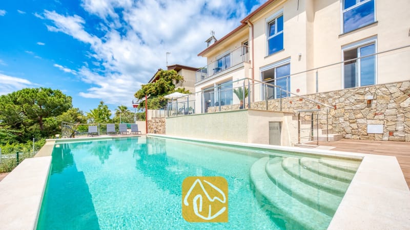 Vakantievilla's Costa Brava Spanje - Villa BellaVista - Groot buitenzwembad met inlooptrap bij Villa BellaVista. De met natuursteen beklede gevel wordt gecombineerd met terrassen voorzien van glazen balustrades, houten vlonders en grijze ligbedden, omringd door weelderig groen.