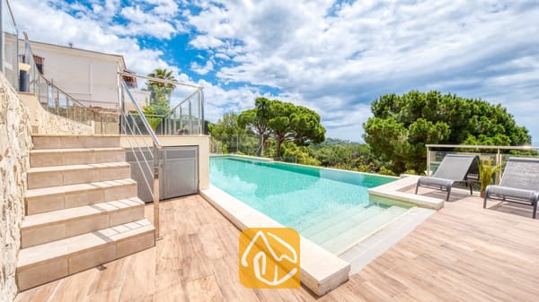 Vakantievilla's Costa Brava Spanje - Villa BellaVista - Privézwembad bij Villa BellaVista met een terras van houtlooktegels, grijze ligbedden en glazen balustrades die uitkijken over groene bomen onder een bewolkte blauwe lucht.