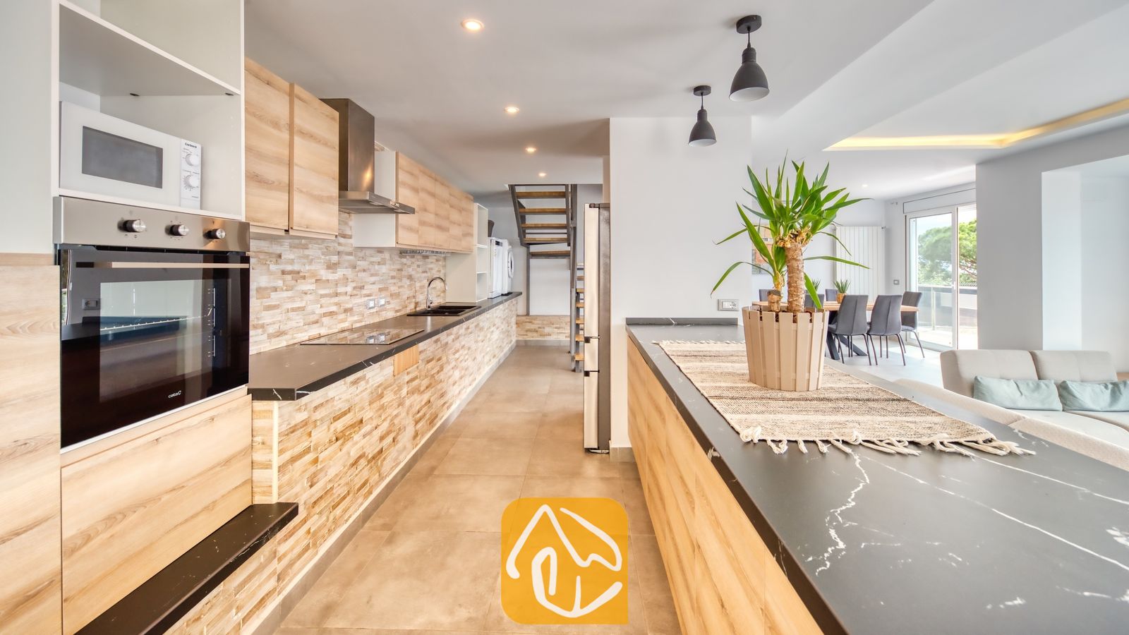 Vakantievilla's Costa Brava Spanje - Villa BellaVista - Moderne keuken in Villa BellaVista met werkbladen in zwart marmerlook, lichthouten kasten en een stenen achterwand. De open indeling sluit naadloos aan op een eethoek met grijze stoelen.