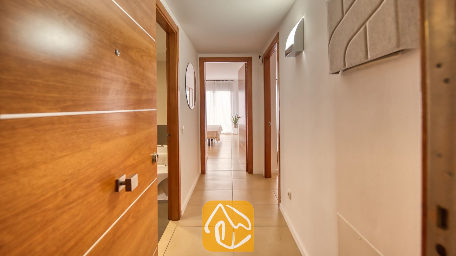 Vakantievilla's Costa Brava Spanje - Apartment Gaudi - De binnenhal van Appartement Gaudi, voorzien van witte muren, tegelvloeren en houten deuren die leiden naar een helder verlichte slaapkamer.