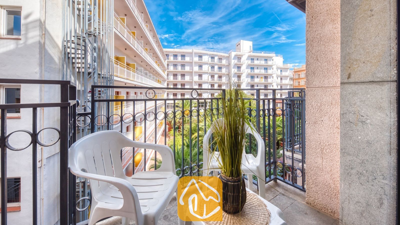 Vakantievilla's Costa Brava Spanje - Apartment Gaudi - Balkon van Appartement Gaudi met twee witte kunststoftoelen, een klein tafeltje met een plant in pot en een metalen balustrade die uitkijkt over de naburige witte gebouwen.