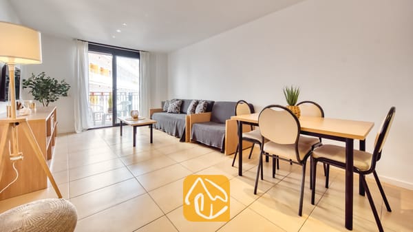Vakantievilla's Costa Brava Spanje - Apartment Gaudi - De woon- en eetkamer van Appartement Gaudi, voorzien van een lichte houten eettafel met vier stoelen, een grijze bank, tegelvloeren en een glazen schuifpui die toegang biedt tot een balkon.