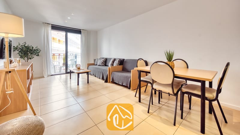 Vakantievilla's Costa Brava Spanje - Apartment Gaudi - De woon- en eetkamer van Appartement Gaudi, voorzien van een lichte houten eettafel met vier stoelen, een grijze bank, tegelvloeren en een glazen schuifpui die toegang biedt tot een balkon.