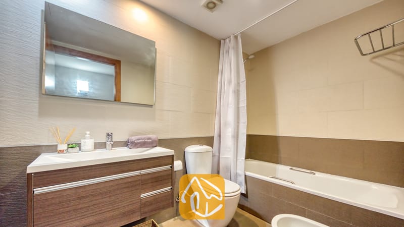 Vakantievilla's Costa Brava Spanje - Apartment Gaudi - Moderne badkamer met een wastafelmeubel in houtlook, een wit toilet en een ligbad met een wit douchegordijn tegen een achtergrond van neutrale beige tegels.