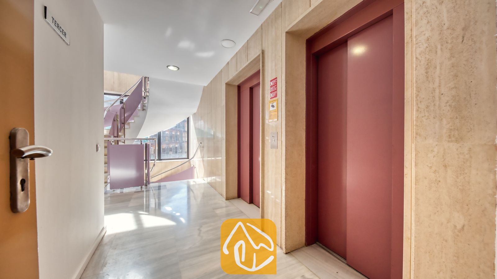 Vakantievilla's Costa Brava Spanje - Apartment Gaudi - Hal van het gebouw met twee rode liftdeuren, een marmeren vloer, beige stenen muren en een gebogen trap met een metalen leuning.
