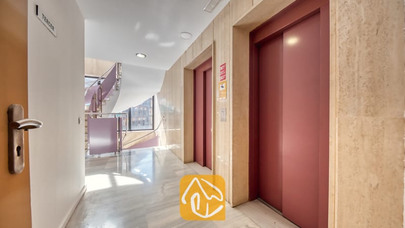 Vakantievilla's Costa Brava Spanje - Apartment Gaudi - Hal van het gebouw met twee rode liftdeuren, een marmeren vloer, beige stenen muren en een gebogen trap met een metalen leuning.