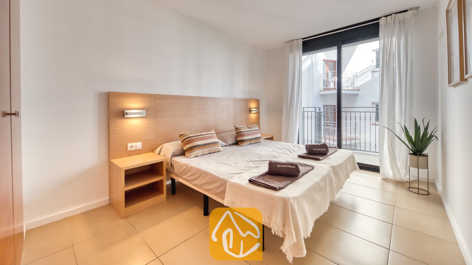 Vakantievilla's Costa Brava Spanje - Apartment Gaudi - Slaapkamer met een tweepersoonsbed, houten hoofdbord met geïntegreerde verlichting, tegelvloeren en een glazen deur die uitkomt op een balkon.