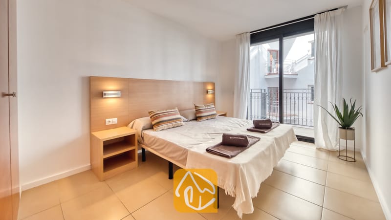 Vakantievilla's Costa Brava Spanje - Apartment Gaudi - Slaapkamer met een tweepersoonsbed, houten hoofdbord met geïntegreerde verlichting, tegelvloeren en een glazen deur die uitkomt op een balkon.