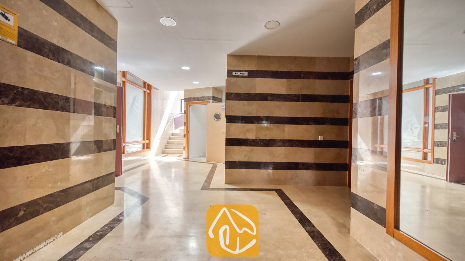 Vakantievilla's Costa Brava Spanje - Apartment Dali - Binnenhal voorzien van beige en donkerbruin gestreepte tegelmuren, lichte marmeren vloeren, inbouwspots in het plafond en een deuropening met de aanduiding BAIXOS in Appartement Dali.