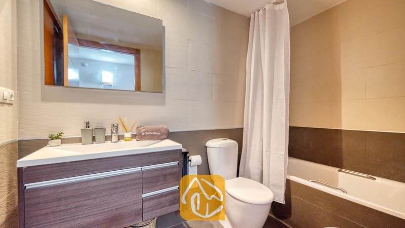 Vakantievilla's Costa Brava Spanje - Apartment Dali - Moderne badkamer met een wastafelmeubel van donker hout met een witte wasbak, een toilet en een ligbad met een wit douchegordijn tegen betegelde muren.