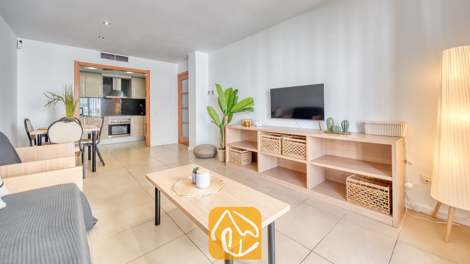 Vakantievilla's Costa Brava Spanje - Apartment Dali - Woonkamer van Appartement Dali met een lichte houten sidetable, een wandtelevisie, rieten opbergmanden en een open keuken op de achtergrond.