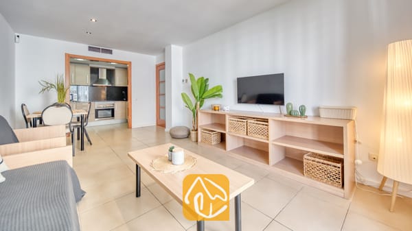 Vakantievilla's Costa Brava Spanje - Apartment Dali - Woonkamer van Appartement Dali met een lichte houten sidetable, een wandtelevisie, rieten opbergmanden en een open keuken op de achtergrond.