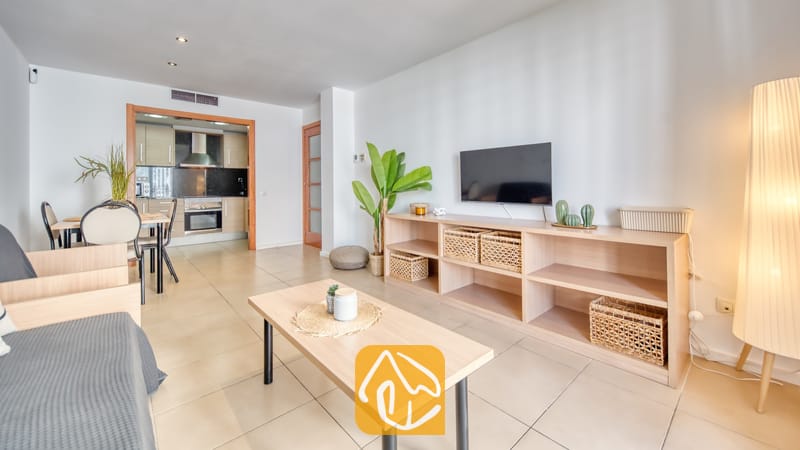 Vakantievilla's Costa Brava Spanje - Apartment Dali - Woonkamer van Appartement Dali met een lichte houten sidetable, een wandtelevisie, rieten opbergmanden en een open keuken op de achtergrond.