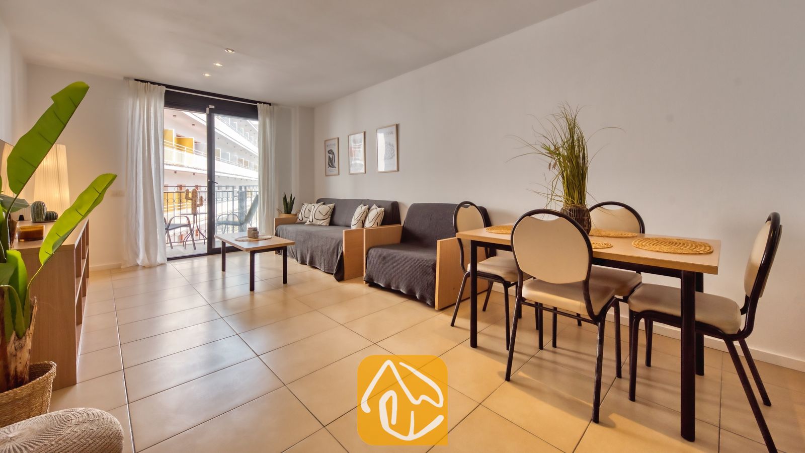 Vakantievilla's Costa Brava Spanje - Apartment Dali - Woon- en eetgedeelte in Appartement Dali, voorzien van een houten tafel met vier stoelen, grijze banken, tegelvloeren en een glazen balkondeur.