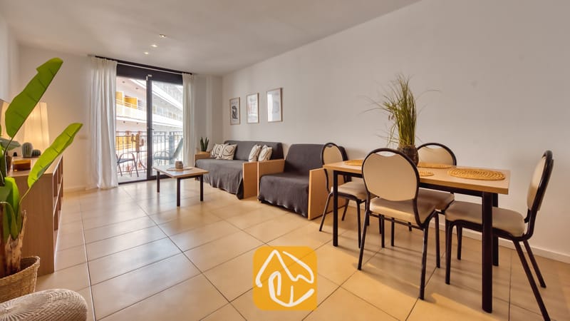 Vakantievilla's Costa Brava Spanje - Apartment Dali - Woon- en eetgedeelte in Appartement Dali, voorzien van een houten tafel met vier stoelen, grijze banken, tegelvloeren en een glazen balkondeur.