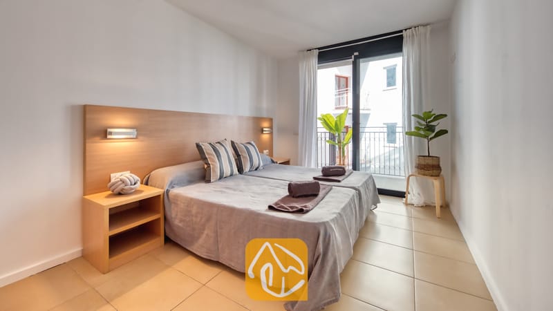Vakantievilla's Costa Brava Spanje - Apartment Dali - Slaapkamer in Appartement Dali met een tweepersoonsbed voorzien van grijs beddengoed, gestreepte kussens en een houten hoofdbord met ingebouwde verlichting dat toegang biedt tot een balkon.