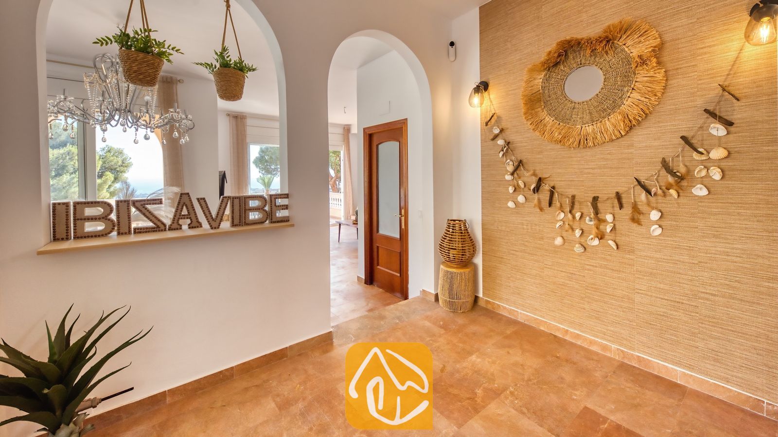 Vakantievilla's Costa Brava Spanje - Villa Ibiza - Interne gang van Villa Ibiza met boogvormige doorgangen, een gestructureerde strokleuren accentmuur gedecoreerd met een schelpenguirlande en een strogen spiegel, en tegelvloeren.