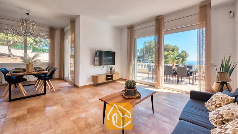 Vakantievilla's Costa Brava Spanje - Villa Ibiza - Ruime woon- en eetkamer in Villa Ibiza met een donkere bank, een houten salontafel met een hangplant of cactus, een wall-mounted televisie en grote glazen schuifdeuren die uitkomen op een zonnig terras met