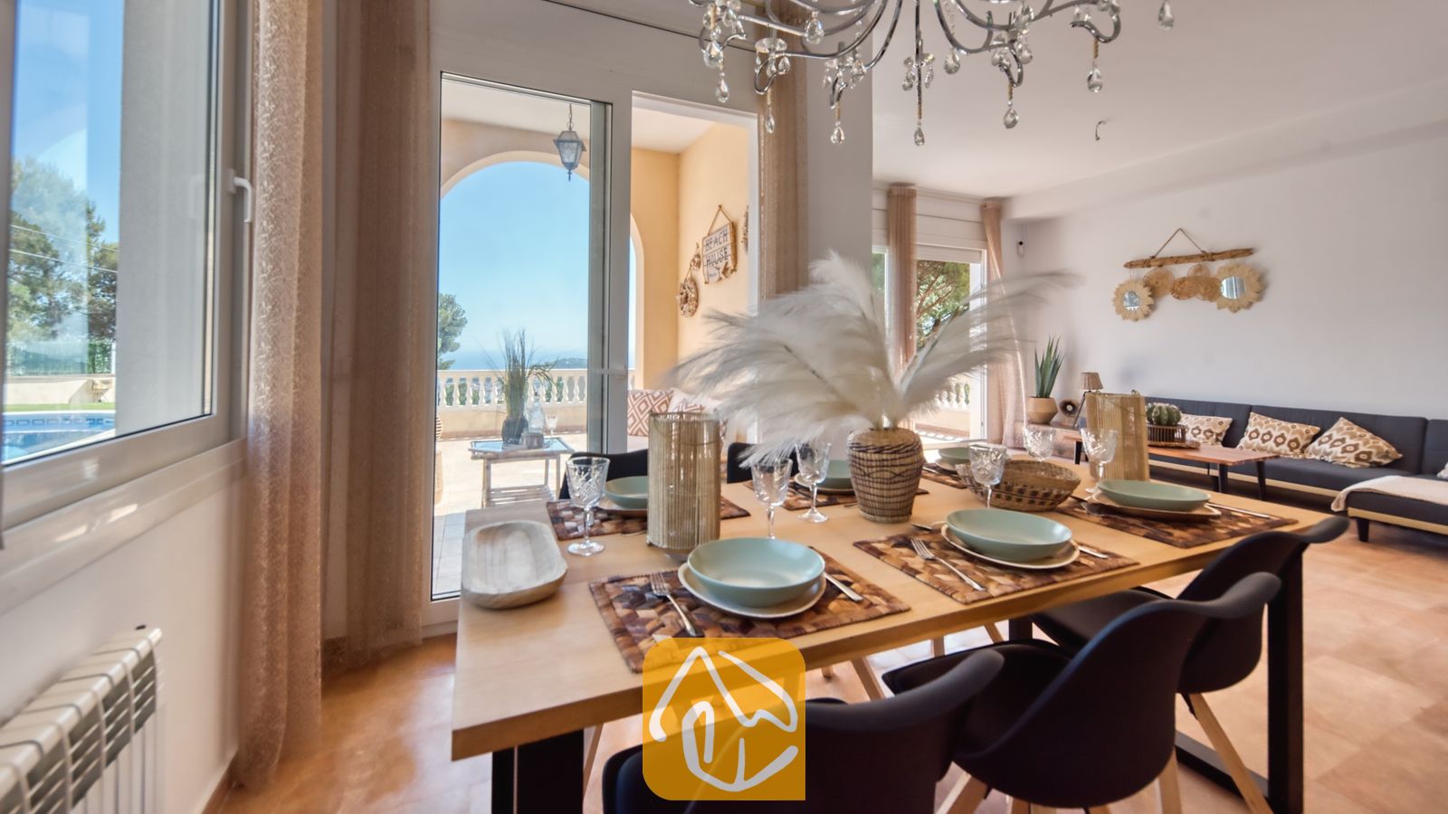 Vakantievilla's Costa Brava Spanje - Villa Ibiza - Houten eettafel met servies, glazen en een groot middelpunt van veren, met glazen deuren die uitkomen op een zonnig terras bij Villa Ibiza.