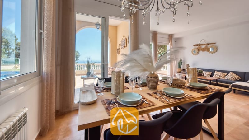 Vakantievilla's Costa Brava Spanje - Villa Ibiza - Houten eettafel met servies, glazen en een groot middelpunt van veren, met glazen deuren die uitkomen op een zonnig terras bij Villa Ibiza.
