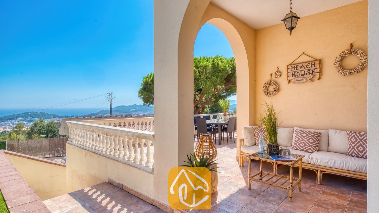 Vakantievilla's Costa Brava Spanje - Villa Ibiza - Overdekt terras bij Villa Ibiza met een beige bank, sierkussens met patronen, een houten salontafel en een boog die uitzicht biedt op de zitplek buiten en het landschap in de verte.