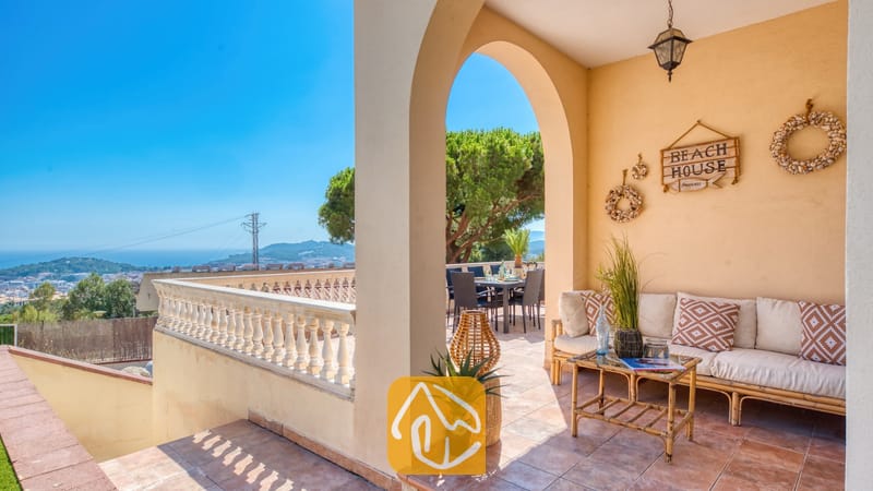 Vakantievilla's Costa Brava Spanje - Villa Ibiza - Overdekt terras bij Villa Ibiza met een beige bank, sierkussens met patronen, een houten salontafel en een boog die uitzicht biedt op de zitplek buiten en het landschap in de verte.