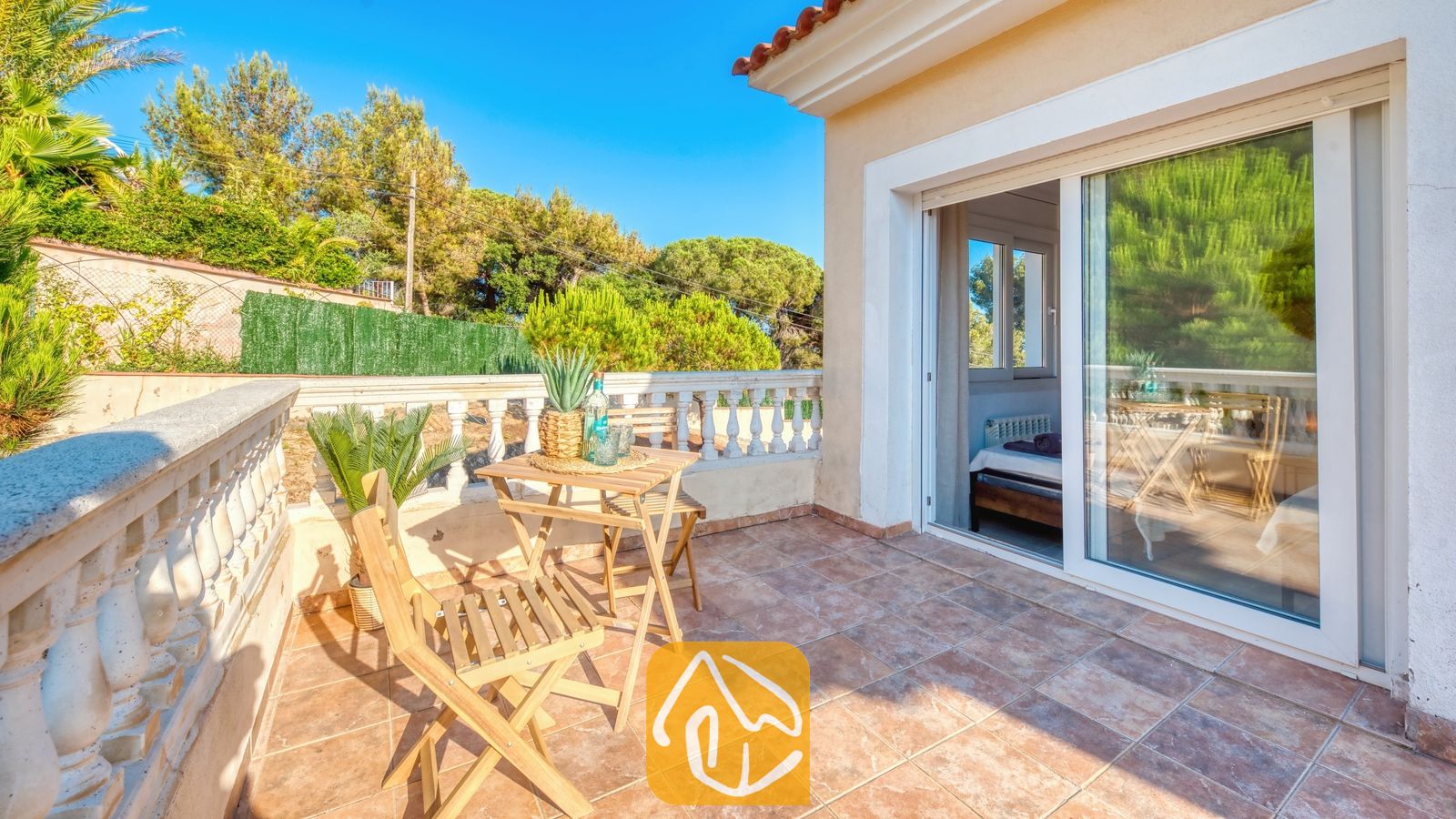 Vakantievilla's Costa Brava Spanje - Villa Ibiza - Zonnig terras bij Villa Ibiza, voorzien van een houten tafel en twee klapstoelen, betegelde vloer, balustrade en glazen schuifpui die uitzicht biedt op het interieur van een slaapkamer.