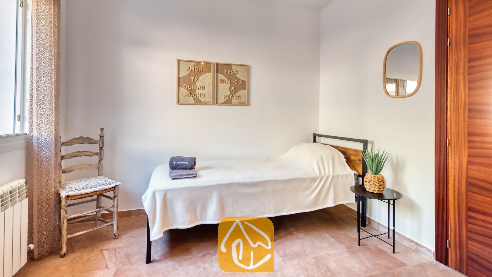 Vakantievilla's Costa Brava Spanje - Villa Ibiza - Een eenpersoonsbed met wit beddengoed en een opgevouwen grijze handdoek, naast een klein zwart bijettafeltje en een rustieke houten stoel in Villa Ibiza.