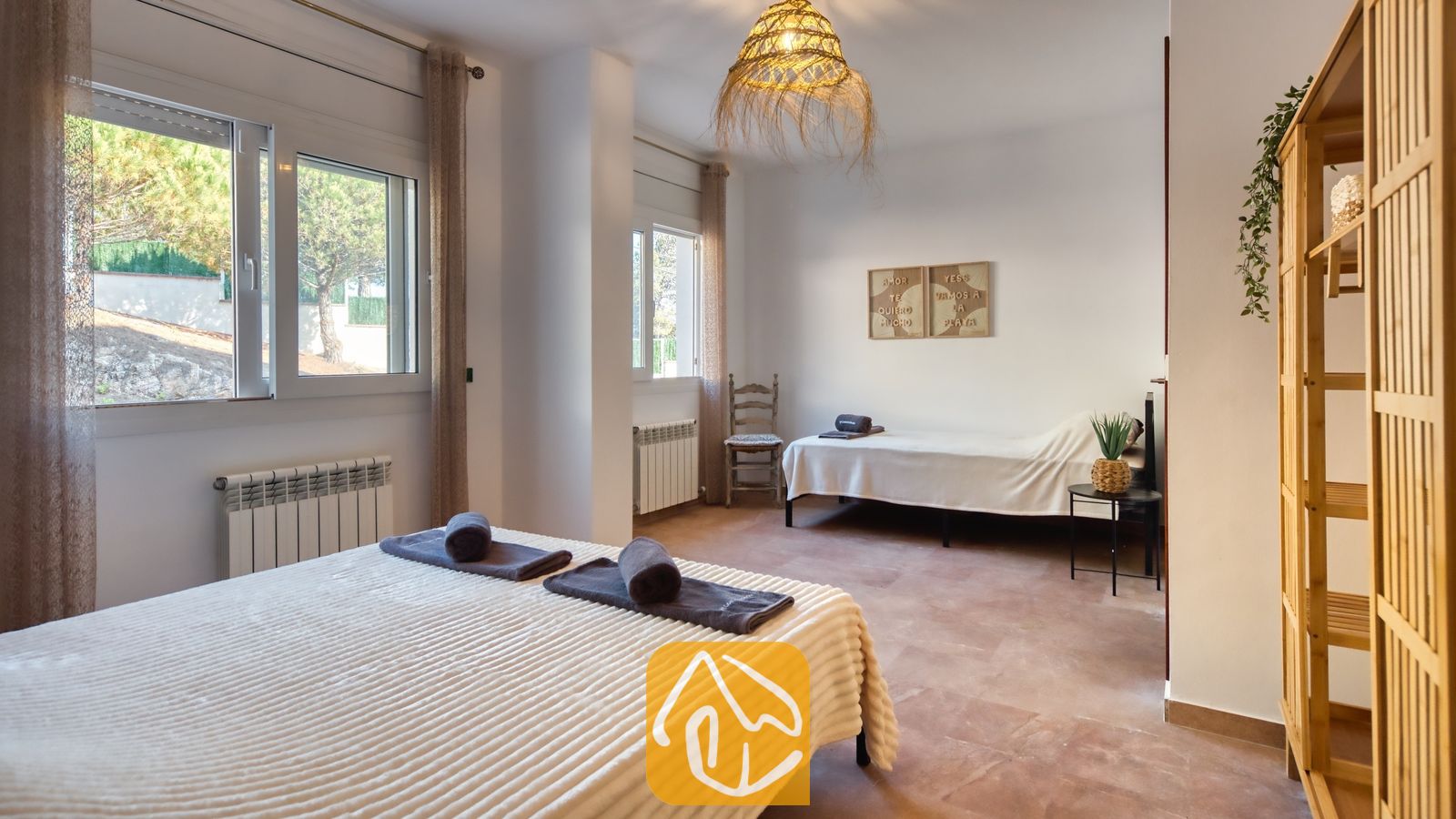 Vakantievilla's Costa Brava Spanje - Villa Ibiza - Slaapkamer met twee eenpersoonsbedden, opgerolde donkere handdoeken, witte muren en een geweven rieten hanglamp aan het plafond.