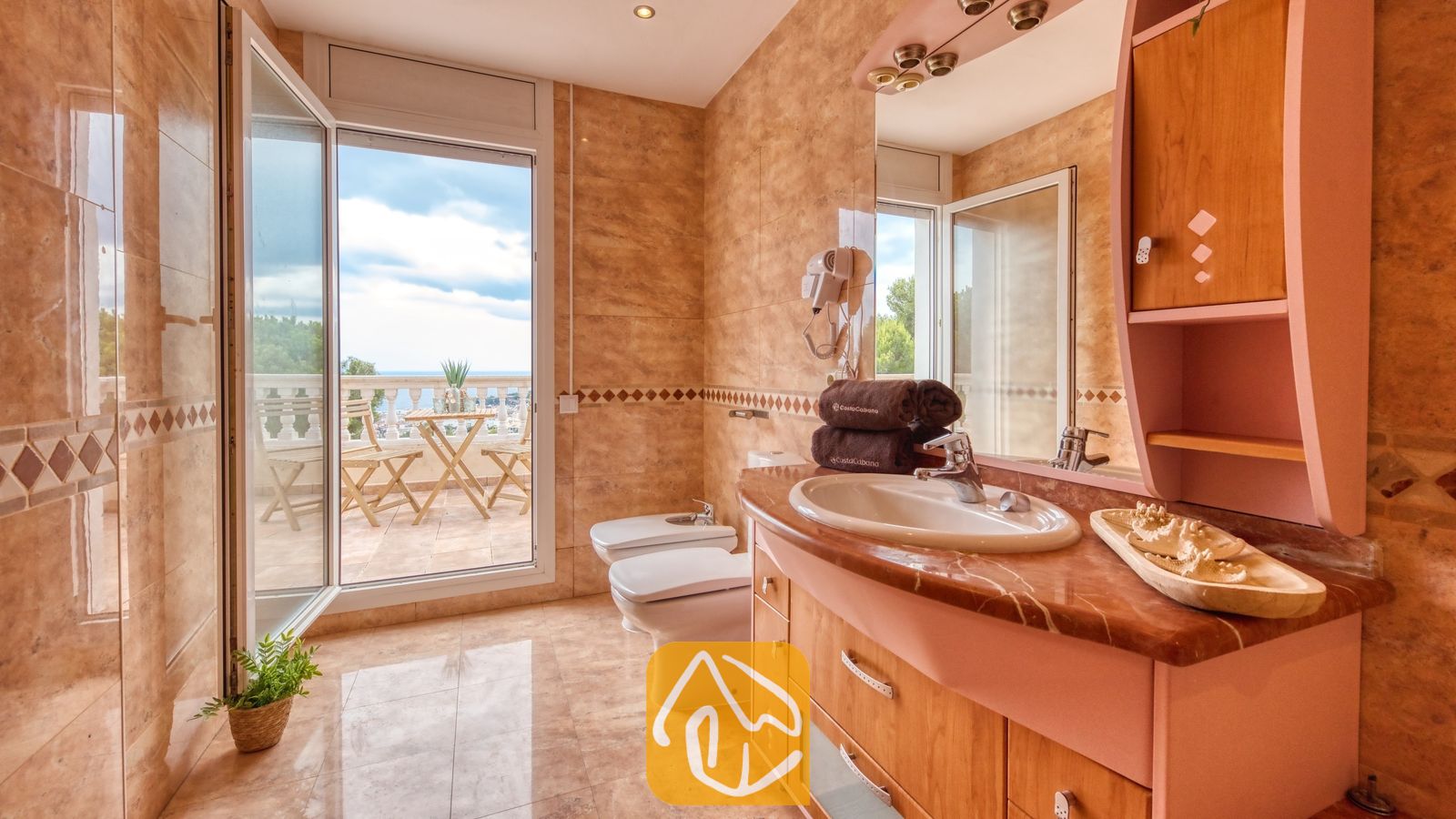 Vakantievilla's Costa Brava Spanje - Villa Ibiza - Badkamer voorzien van wandtegels met marmerlook, een houten badkamermeubel met een ingebouwde wastafel en een open glazen deur die leidt naar een balkon met uitzicht op zee.