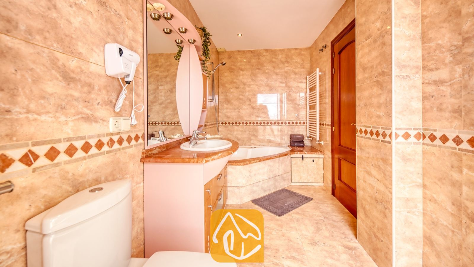 Vakantievilla's Costa Brava Spanje - Villa Ibiza - Badkamer voorzien van beige tegels met marmerlook, een hoekbad en een wastafelmeubel met een roze, boogvormige spiegel.