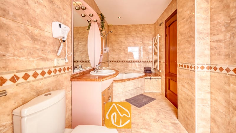 Villas de vacances Costa Brava Espagne - Villa Ibiza - Salle de bains dotée de carreaux effet marbre beige, d'une baignoire d'angle et d'un meuble sous-vasque surmonté d'un miroir rose en arc.