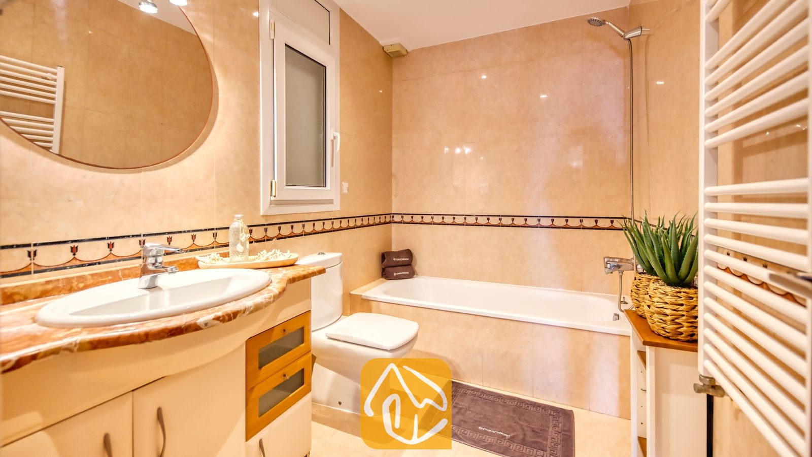 Vakantievilla's Costa Brava Spanje - Villa Ibiza - Badkamer voorzien van wandtegels met een beige marmerlook, een wit keramisch ligbad, een wastafelmeubel met een ronde spiegel en een kamerplant.
