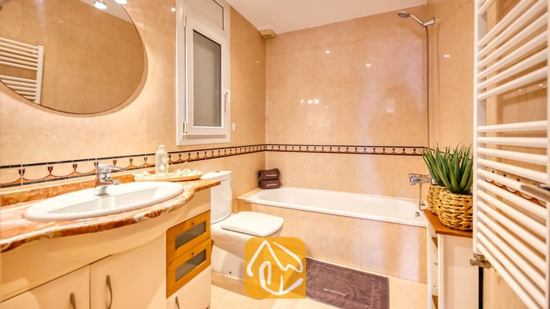 Villas de vacances Costa Brava Espagne - Villa Ibiza - Salle de bains dotée de carrelage mural beige effet marbre, d'une baignoire en céramique blanche, d'un meuble sous-vasque avec un miroir rond et d'une plante en pot.