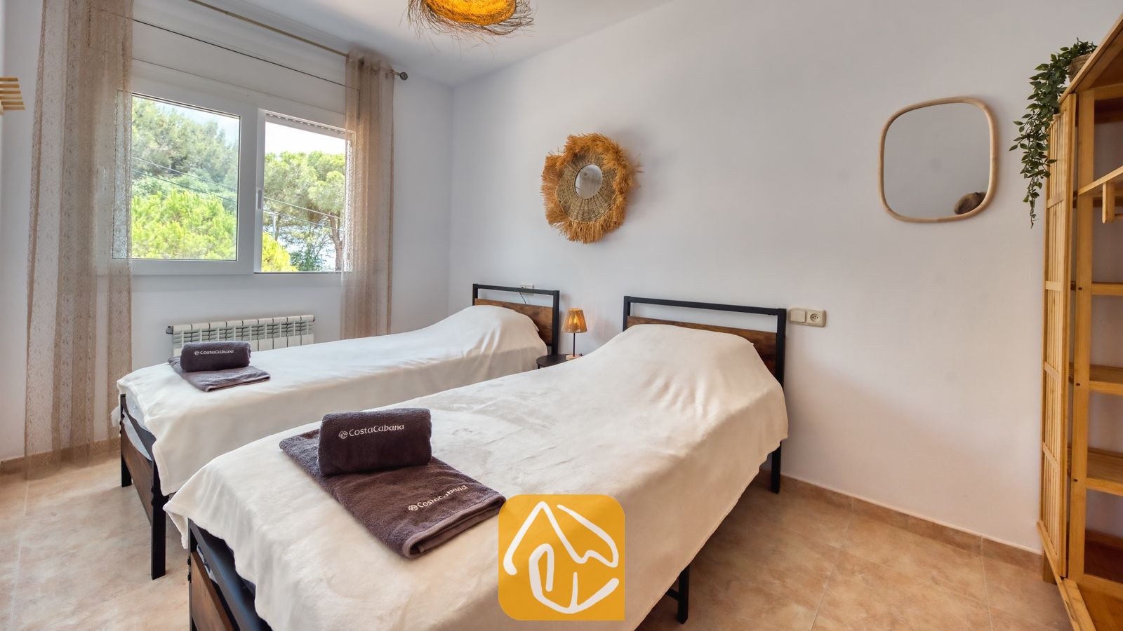 Vakantievilla's Costa Brava Spanje - Villa Ibiza - Slaapkamer met twee eenpersoonsbedden met donkere metalen frames, witter beddengoed, opgevouwen grijze handdoeken en een raam met uitzicht op de bomen.