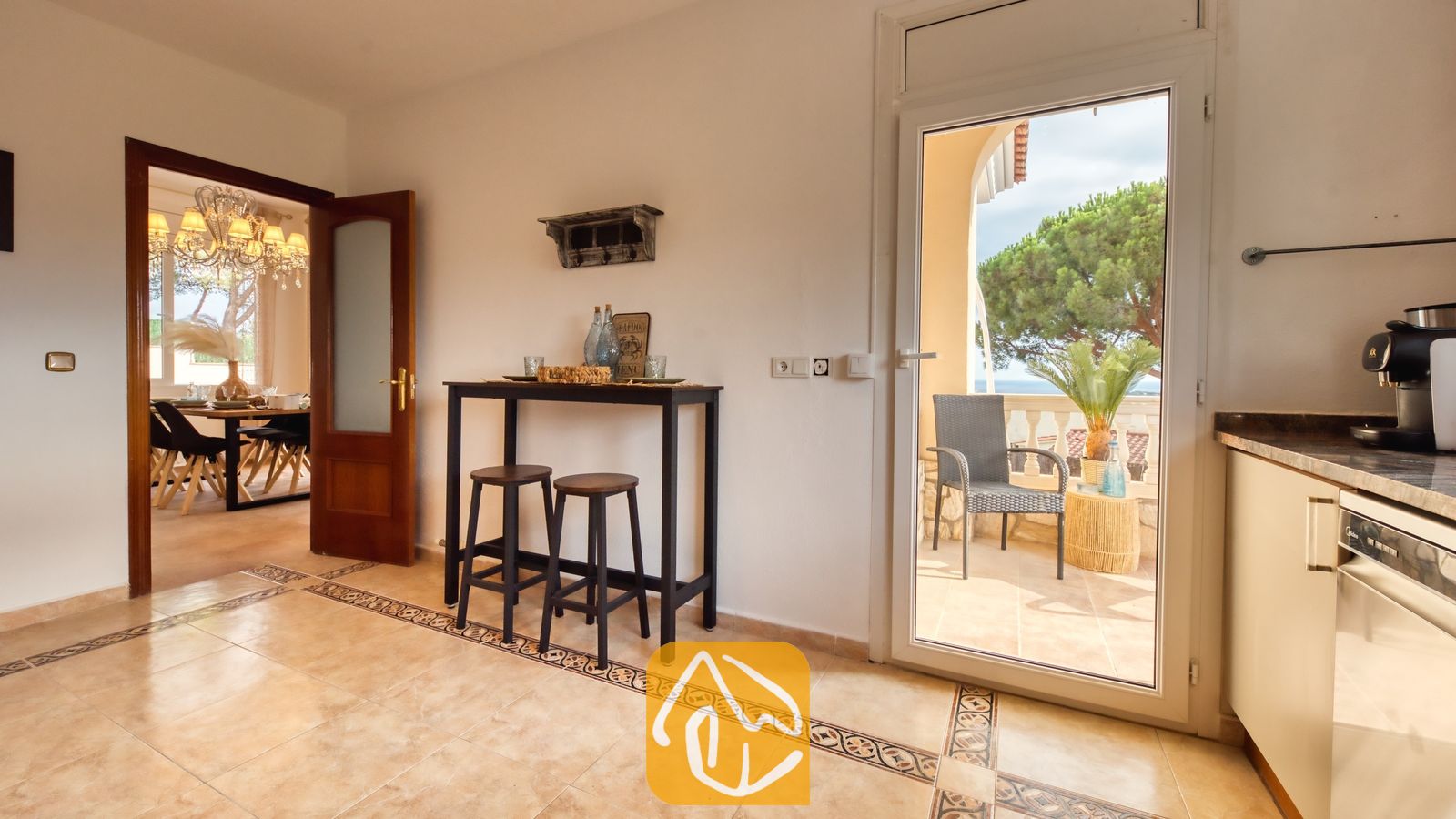 Vakantievilla's Costa Brava Spanje - Villa Ibiza - Interieur van Villa Ibiza met een keukengedeelte voorzien van een hoge bartafel, twee houten krukken, tegelvloeren en een open glazen deur die toegang biedt tot een balkon.