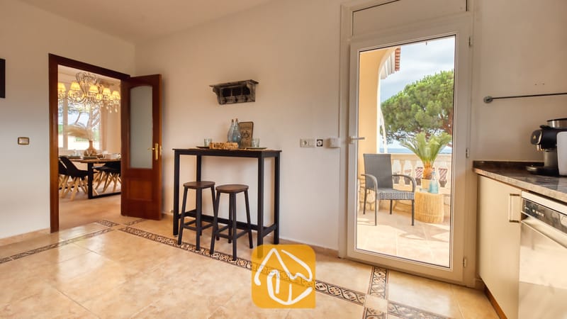 Villas de vacances Costa Brava Espagne - Villa Ibiza - Vue intérieure de la Villa Ibiza montrant un coin cuisine doté d'une console haute, de deux tabourets en bois, de sols carrelés et d'une porte vitrée ouverte donnant sur un balcon.